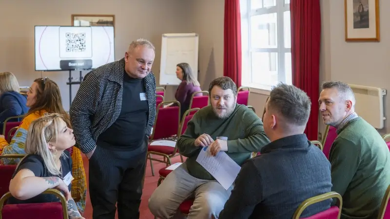 A group of people sitting in a circle in a conference room with red chairs, engaged in discussion. A group of people sitting in a circle in a conference room with red chairs, engaged in discussion.