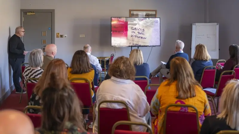 A man presents in front of a seated audience, displaying slides on a large screen. A man presents in front of a seated audience, displaying slides on a large screen. The room is calm and focused, fostering attentive learning.