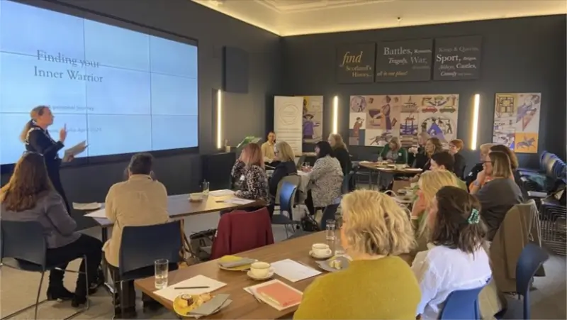 A presenter stands by a large screen displaying "Finding your Inner Warrior" to a room of seated attendees. A presenter stands by a large screen displaying "Finding your Inner Warrior" to a room of seated attendees. The atmosphere is engaged and attentive.