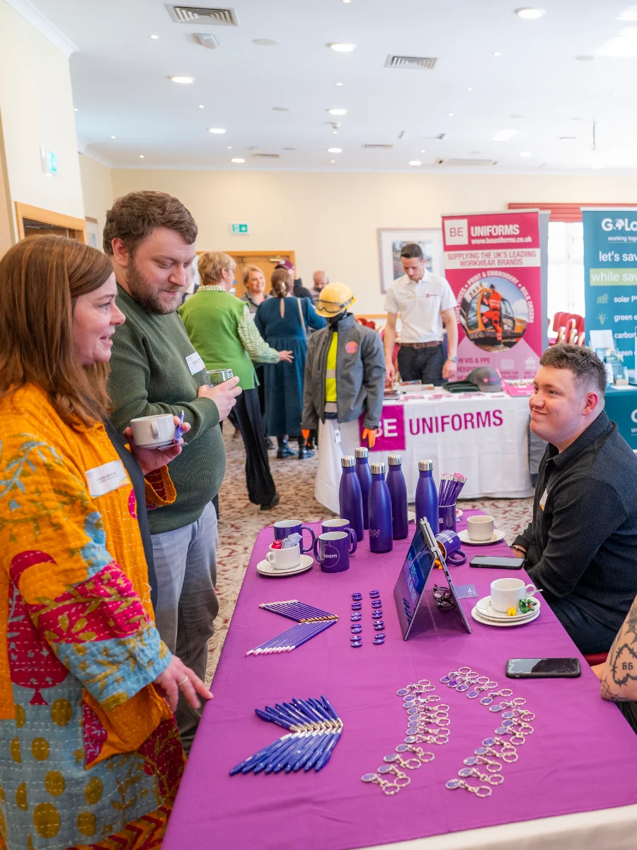 Two people engaging with an exhibitor at a booth with promotional items on a table at a lively networking event.