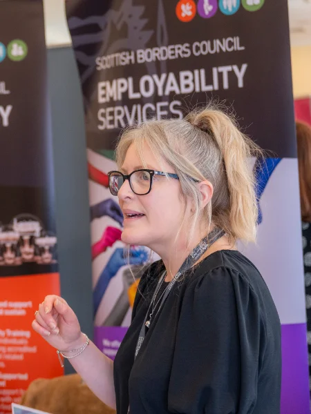A woman with glasses and a ponytail speaks animatedly in front of Scottish Borders Council Employability Services banner