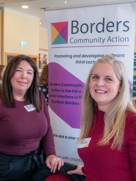 Two women smiling in front of a "Borders Community Action" banner. They wear name tags and the setting is a bright indoor event space.