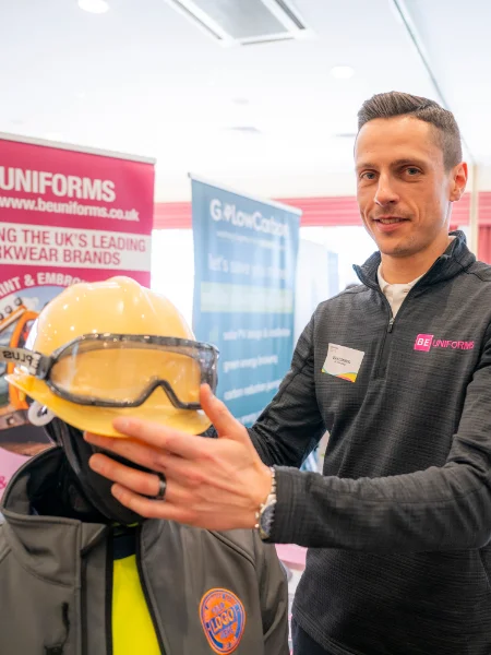 Man in a branded jacket stands beside a mannequin wearing a yellow hard hat and goggles. Background banners indicate a branding event.