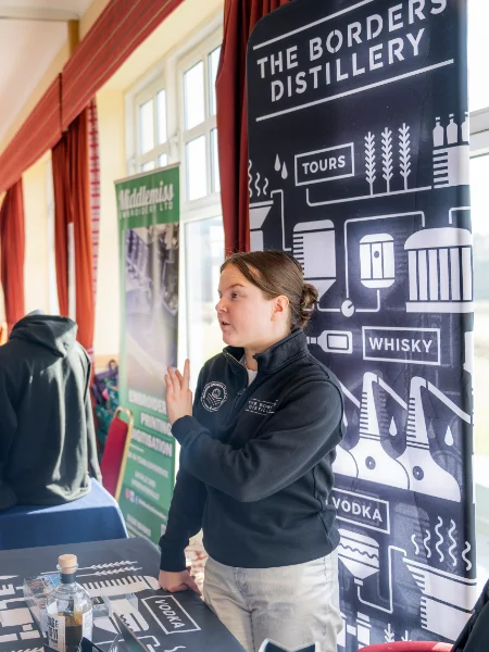 Young woman in a black jacket presents at a distillery booth. Banners display "The Borders Distillery" with icons of whisky and vodka. Bright, engaging scene.