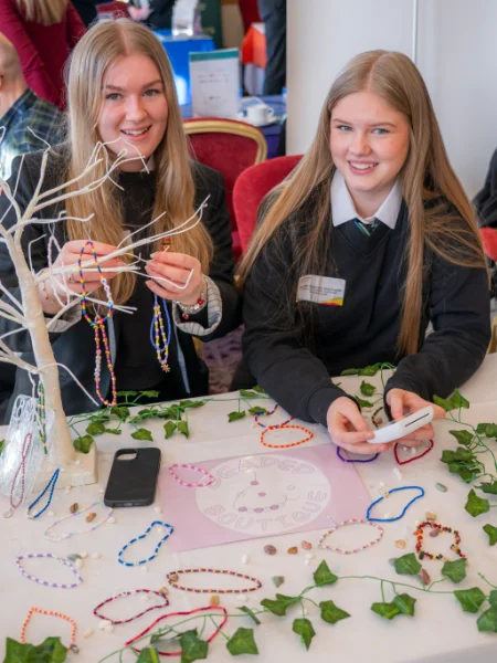 Two school students smile while arranging colourful bead bracelets on a display tree at a table with ivy decor. The mood is cheerful and creative.